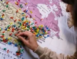 A woman, with only her side profile and hand in view, places a pin on a map of North America which is covered with other pins.