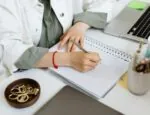 A person working at a desk writing in a spiral notebook. Only the desk and person's hands are visible.