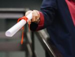A person wearing a graduation gown, with their face out of frame, holding a rolled-up diploma toward the camera.