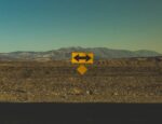 A road sign with arrows pointing left and right in front of a mountain landscape.
